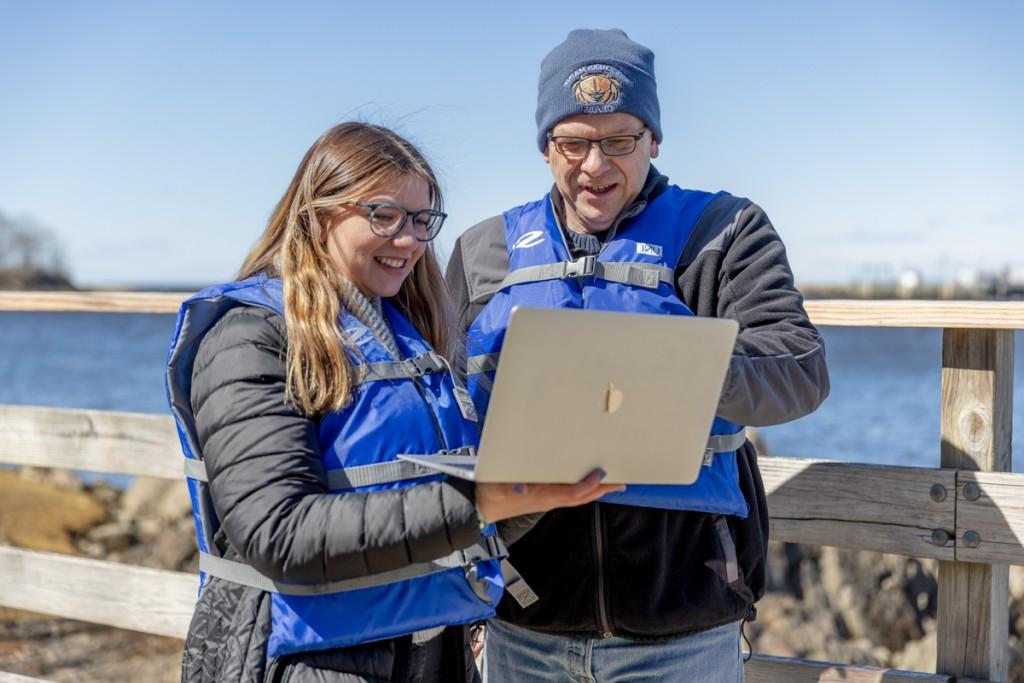 一名学生穿着救生衣站在海边，和马库斯·弗雷德里克教授一起在笔记本电脑上查看数据