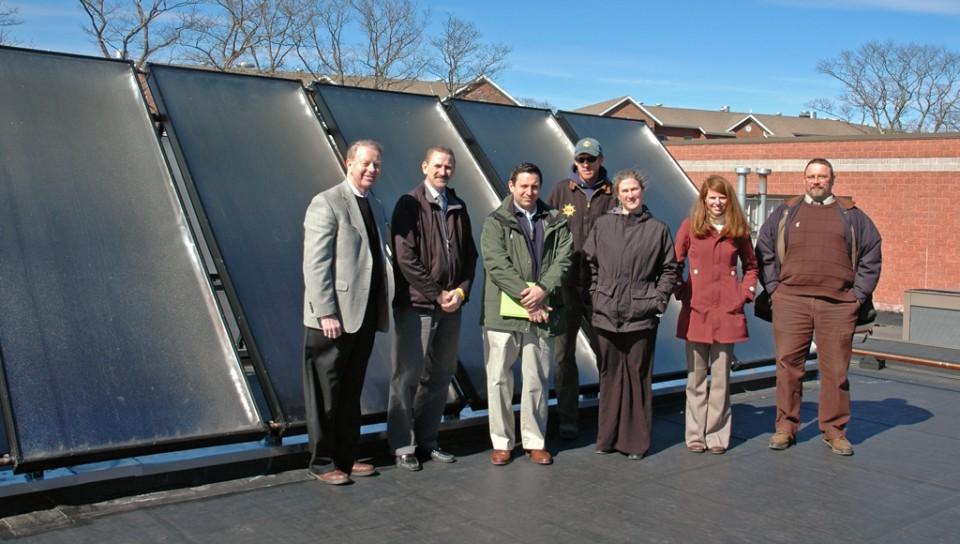 A group of U N E faculty and staff stand in front of five large solar panels