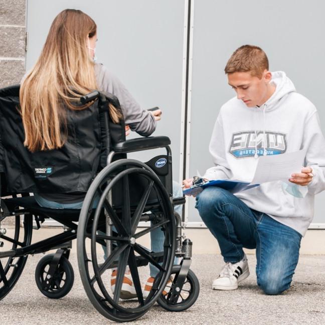 一个拿着剪贴板的学生跪在另一个坐在轮椅上的学生旁边
