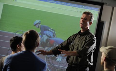正规澳门赌场网络教授吉姆·卡瓦诺(Jim Cavanaugh)在屏幕前讲课，屏幕上可以看到骑自行车的人