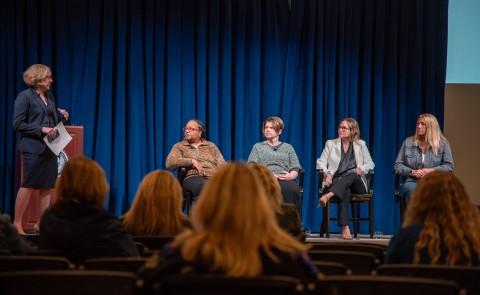 Photo shows five women on stage, four sit in chairs and another stands to moderate a panel discussion